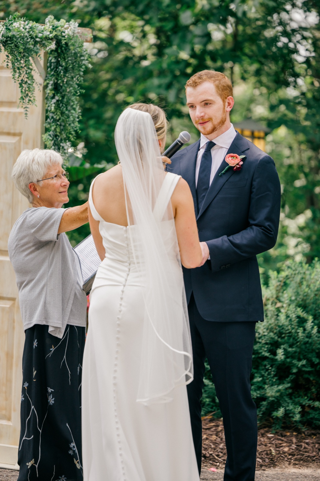 The groom looks intently at his bride during the ceremony as the officiant holds a microphone beneath a greenery arch — Tim Larsen Photography, Brainerd Lakes MN