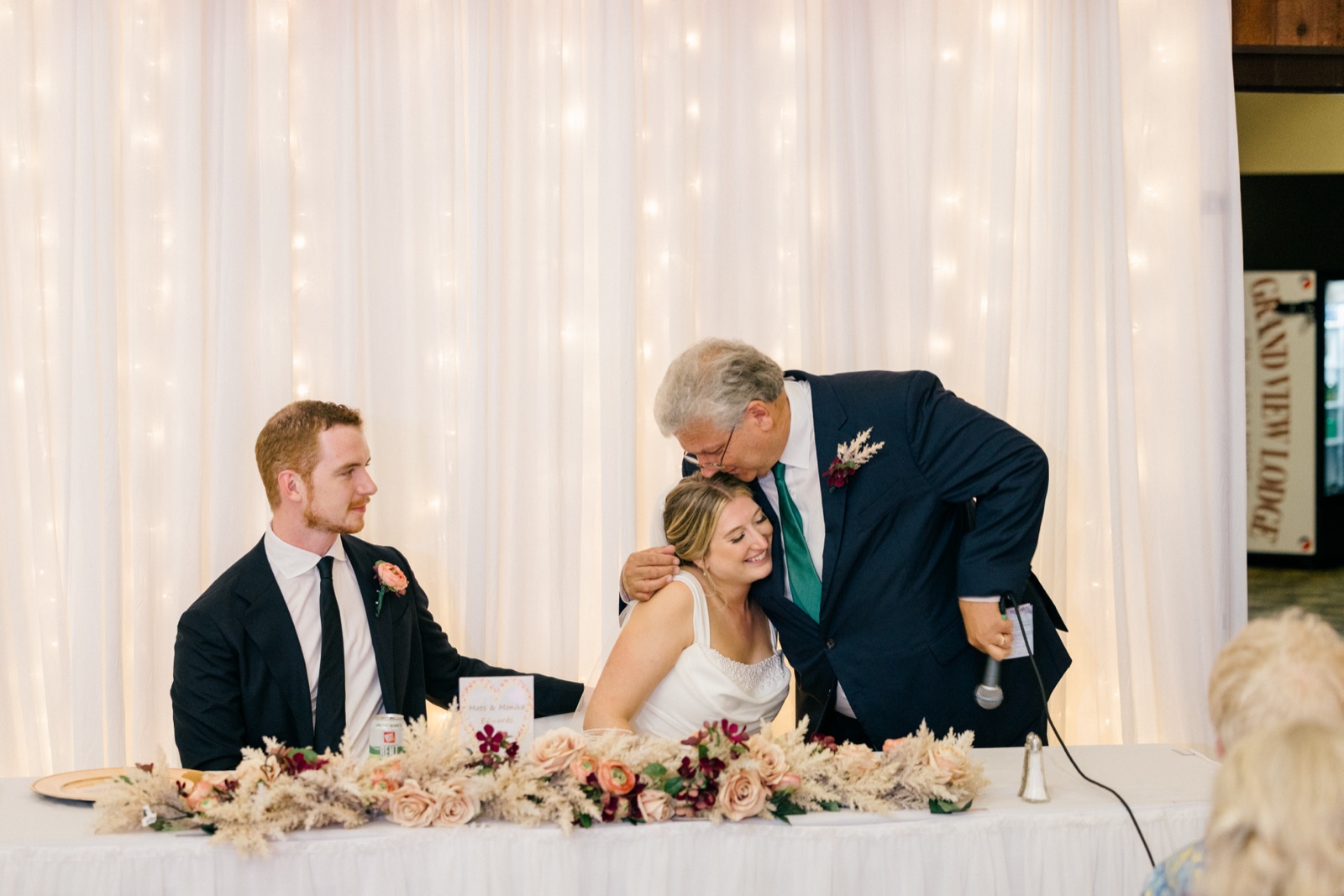 The bride's father leans down to kiss the top of her head at the reception head table while the groom looks on with a soft smile — Tim Larsen Photography, Brainerd Lakes MN