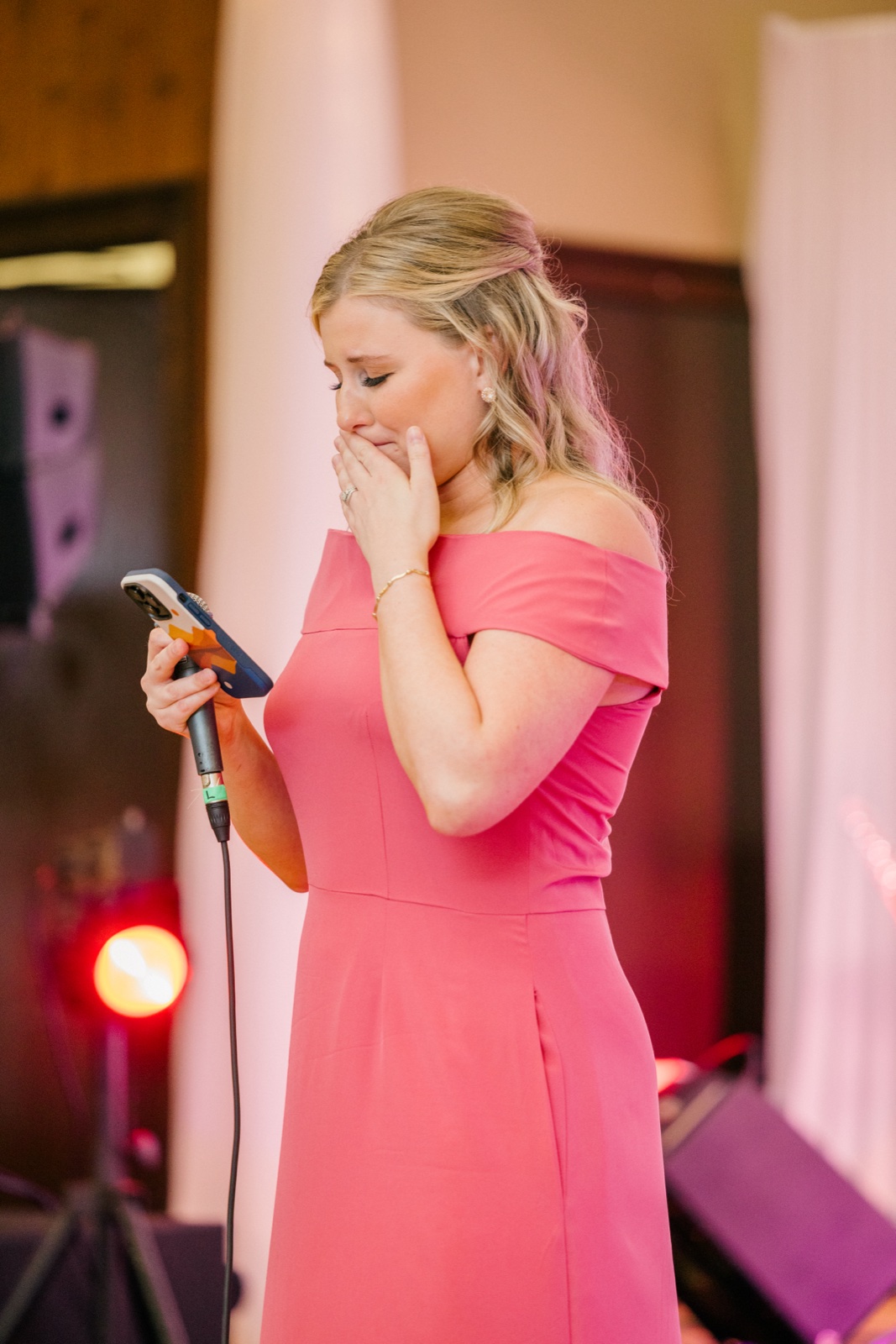 A bridesmaid in a pink off-shoulder dress covers her mouth, overcome with emotion mid-toast as she holds a phone and microphone — Tim Larsen Photography, Brainerd Lakes MN
