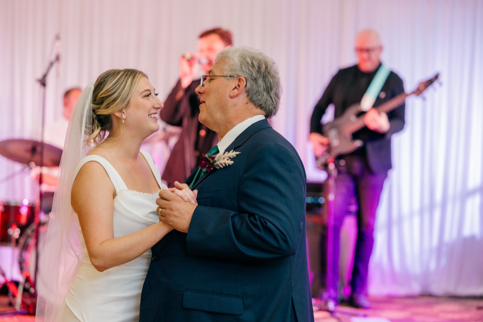 Monika smiles at her father as they dance together, with a live band playing softly behind them on stage — Tim Larsen Photography, Brainerd Lakes MN