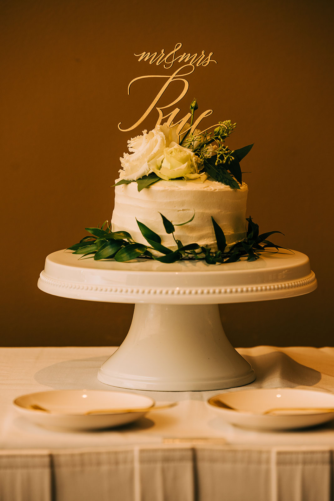 Wedding cake with gold calligraphy topper — white frosting, greenery, and white roses — Tim Larsen Photography, Brainerd Lakes MN