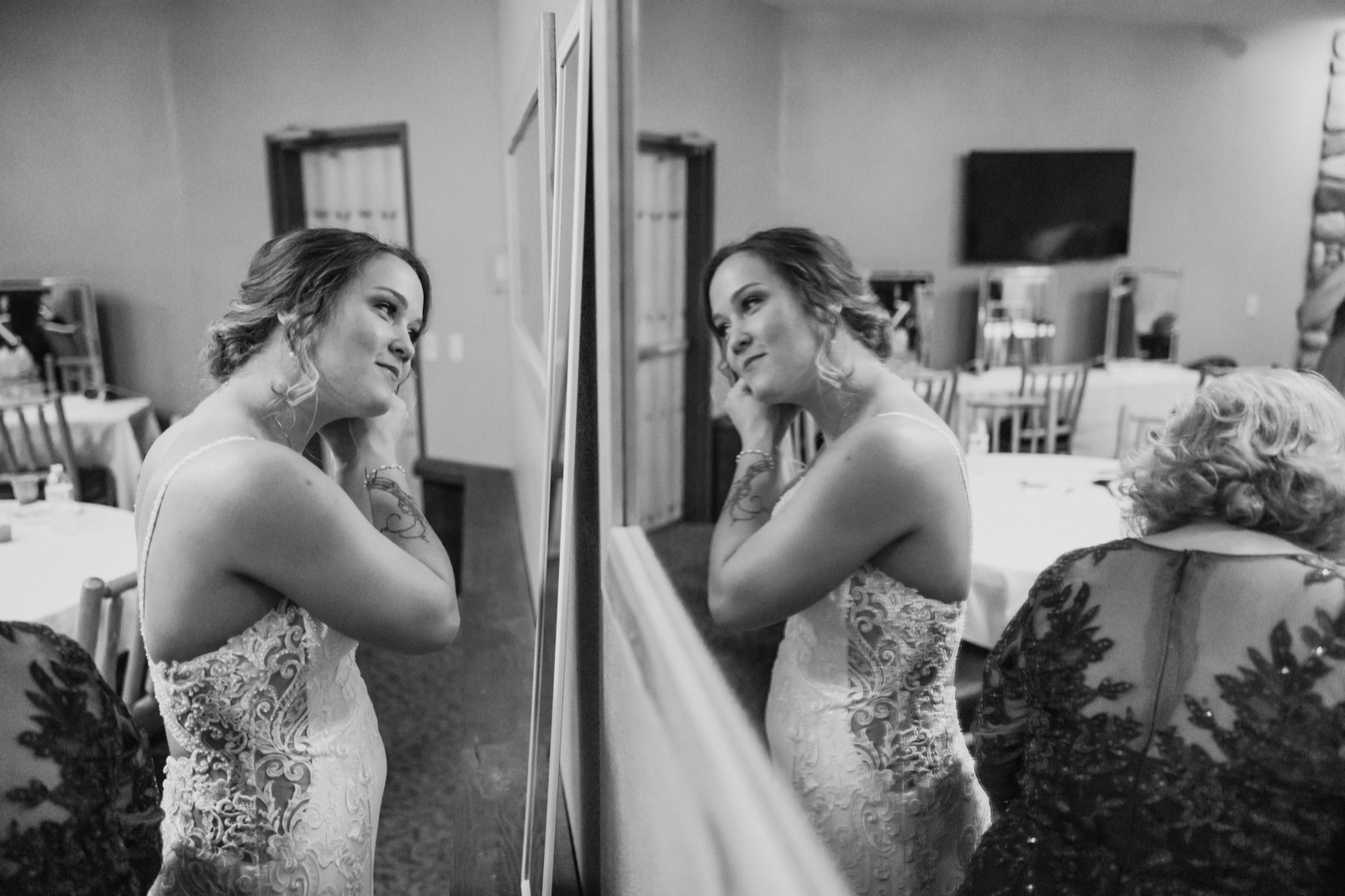 The bride fastens an earring while looking at her reflection in a full-length mirror, her mother visible in the foreground in a lace dress — Tim Larsen Photography, Brainerd Lakes MN