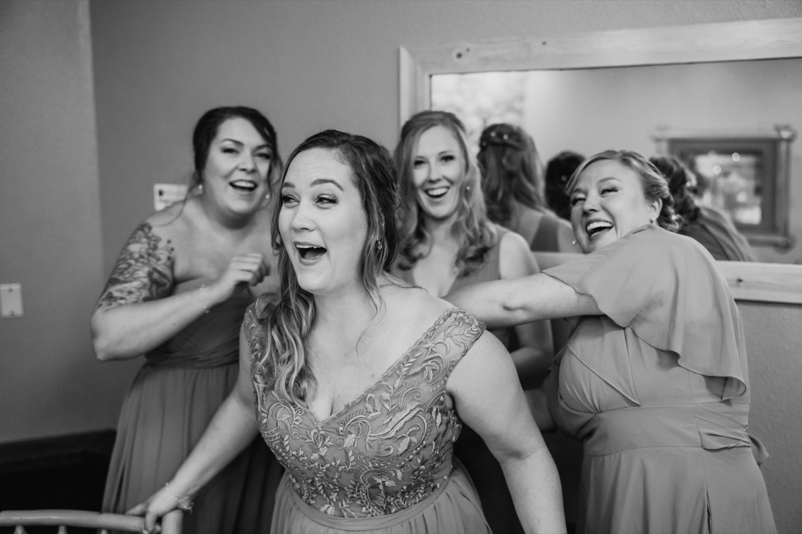 The bride laughs open-mouthed in a lace gown while three bridesmaids in matching dresses laugh around her in a Whitefish Lodge bridal suite — Tim Larsen Photography, Brainerd Lakes MN