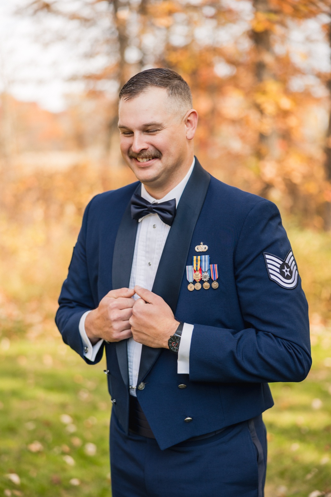 The groom in a navy military dress uniform with service medals laughs while buttoning his jacket against a backdrop of autumn foliage — Tim Larsen Photography, Brainerd Lakes MN