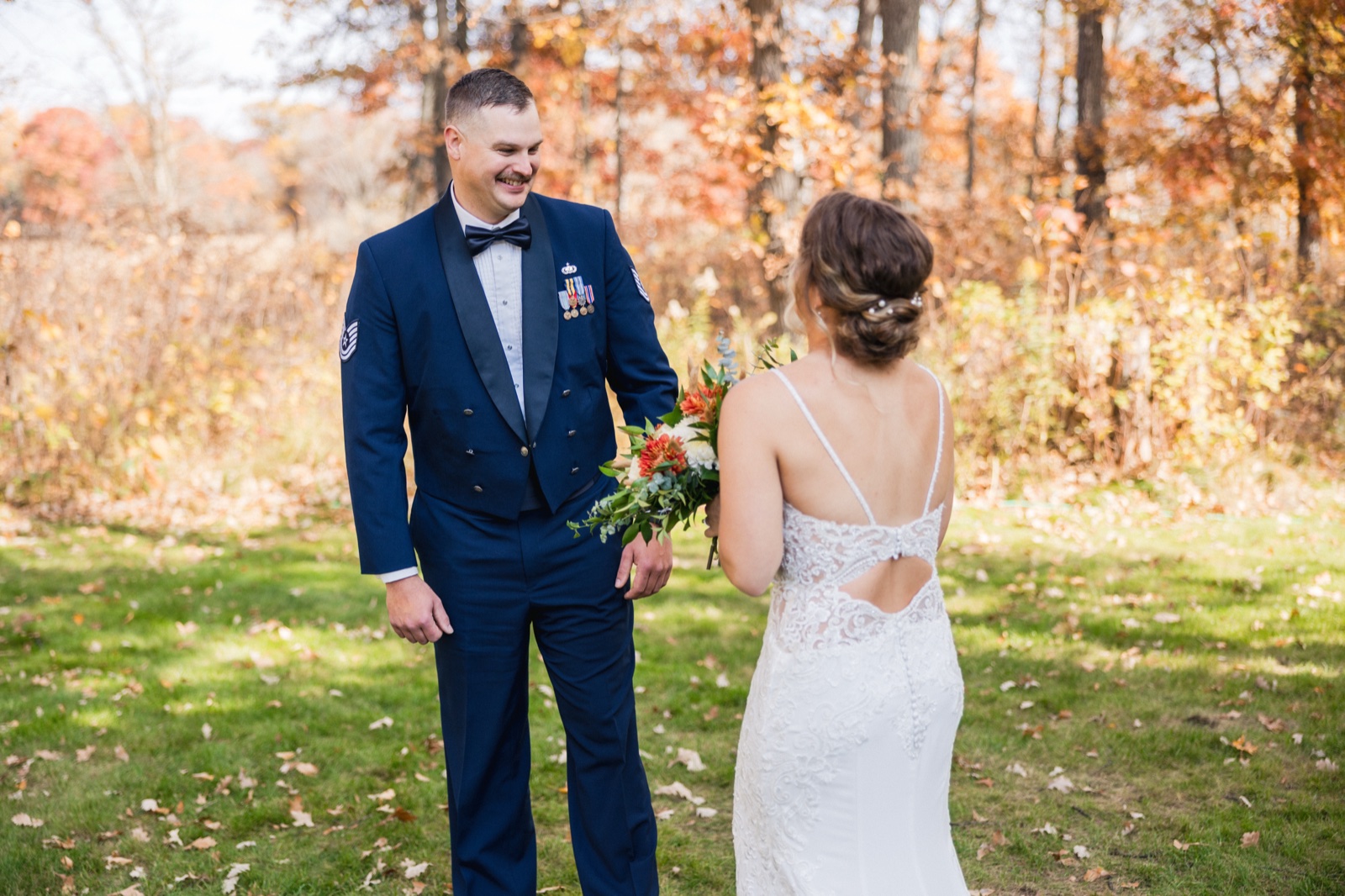 The groom in a military dress uniform smiles at the bride, who stands with her back to the camera holding a bouquet, surrounded by autumn trees — Tim Larsen Photography, Brainerd Lakes MN