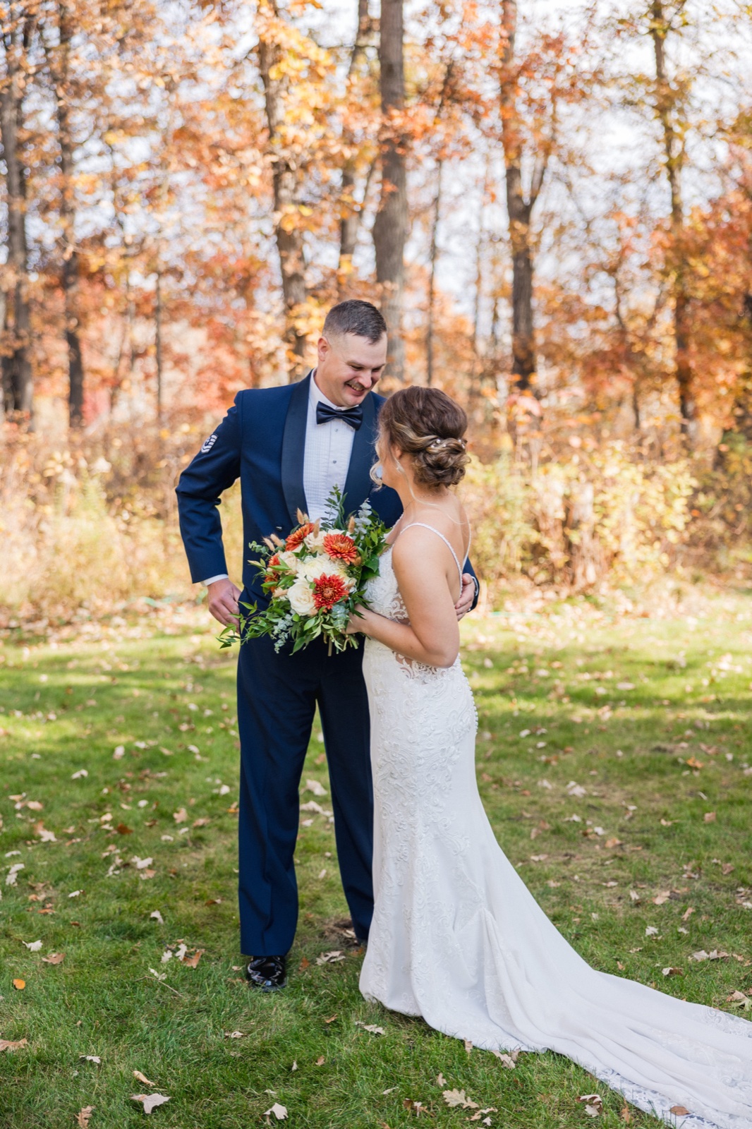 The bride and groom stand close together on a grassy lawn, smiling at each other as she holds a bouquet of orange and cream dahlias against a backdrop of autumn trees — Tim Larsen Photography, Brainerd Lakes MN