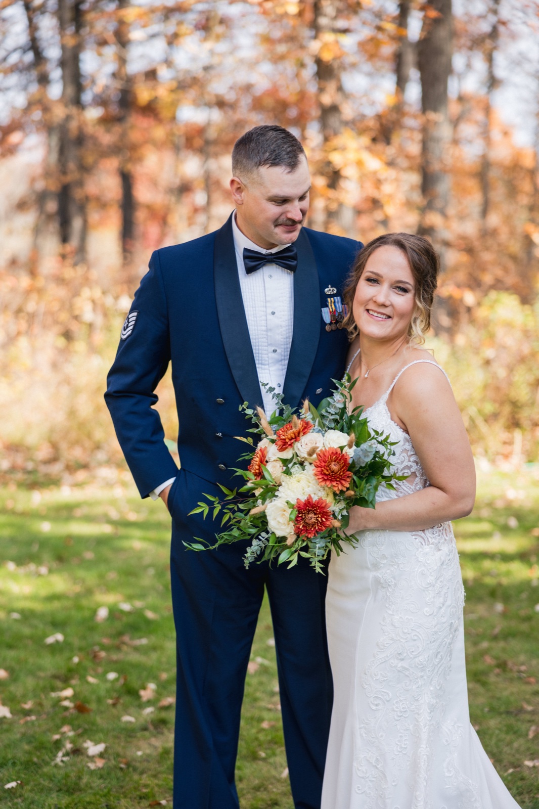 The groom in a military dress uniform looks down at the bride, who smiles toward the camera while holding a fall bouquet of orange and cream blooms in a wooded autumn setting — Tim Larsen Photography, Brainerd Lakes MN