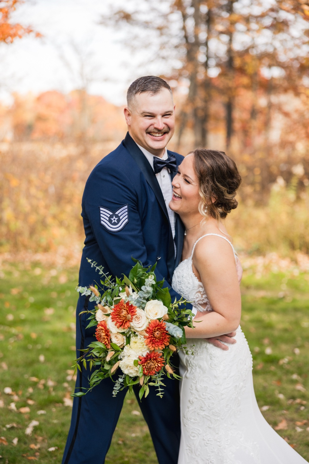 The groom in a military dress uniform grins broadly while the bride leans into him laughing, her bouquet of orange and cream blooms held to the side, against a backdrop of autumn trees — Tim Larsen Photography, Brainerd Lakes MN
