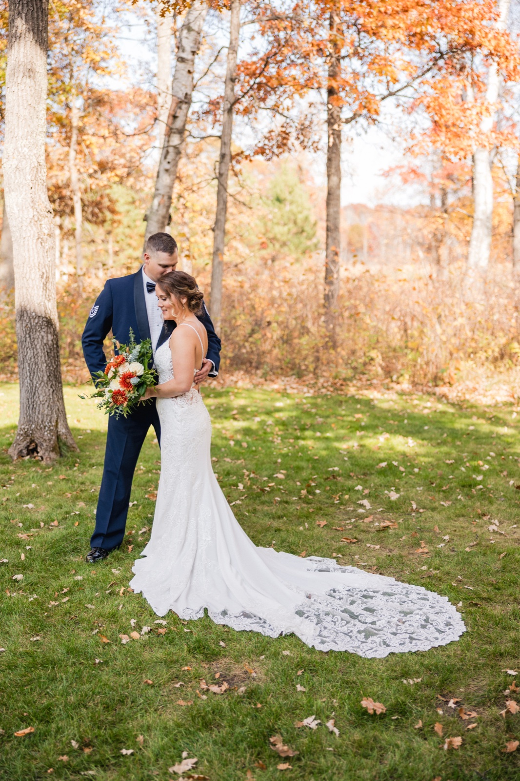 The groom in a military dress uniform kisses the bride's forehead as she holds a fall-toned bouquet, her lace train spread across the grass beneath autumn birch trees — Tim Larsen Photography, Brainerd Lakes MN