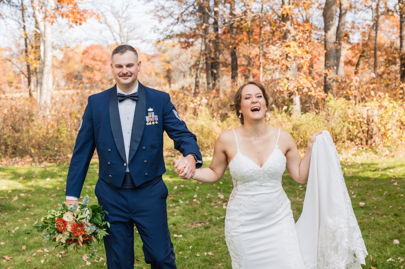 The bride laughs openly while walking hand-in-hand with her groom in military dress uniform across an autumn lawn, her veil lifted in her free hand — Tim Larsen Photography, Brainerd Lakes MN