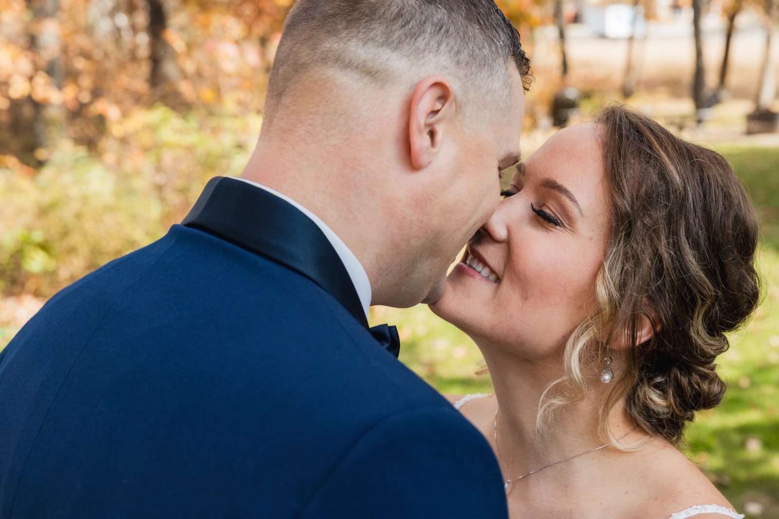 The groom leans in close to the bride's face as she smiles wide, photographed from behind his shoulder against an autumn-toned background — Tim Larsen Photography, Brainerd Lakes MN