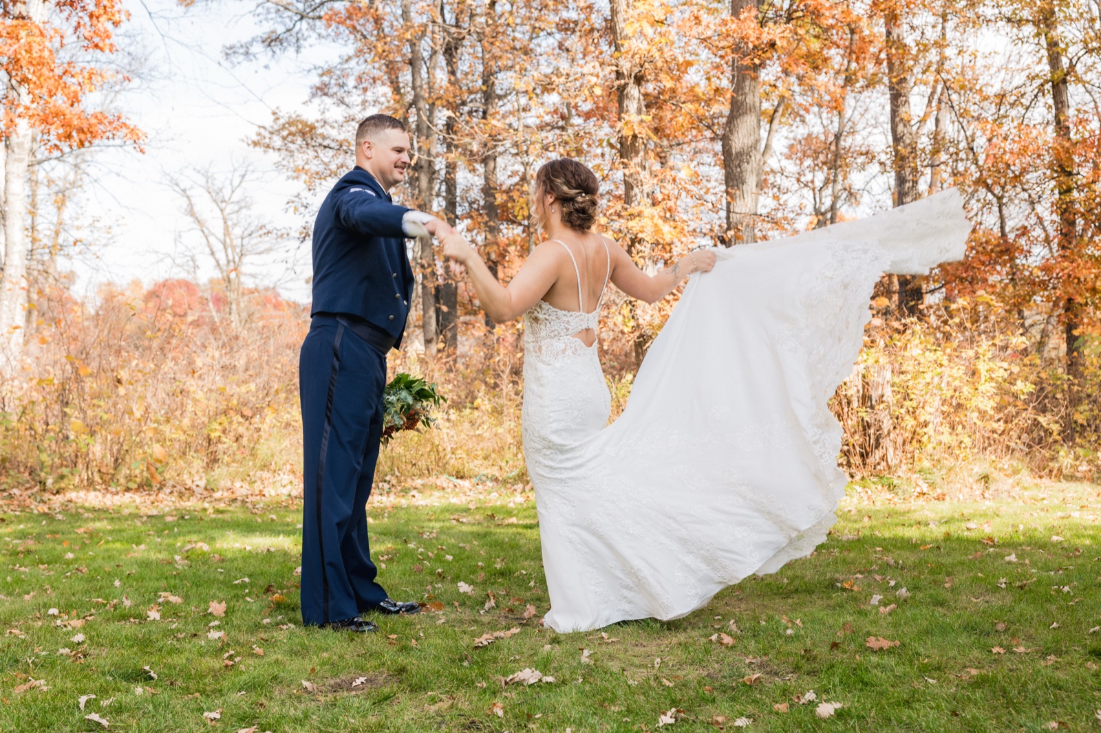 The groom in dress blues twirls the bride as her lace train fans out across a wooded clearing in fall color — Tim Larsen Photography, Brainerd Lakes MN