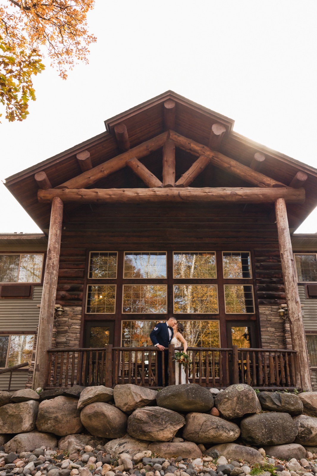 The bride and groom share a kiss on the timber-framed porch of Whitefish Lodge, dwarfed by the lodge's massive log gable and stone foundation — Tim Larsen Photography, Brainerd Lakes MN