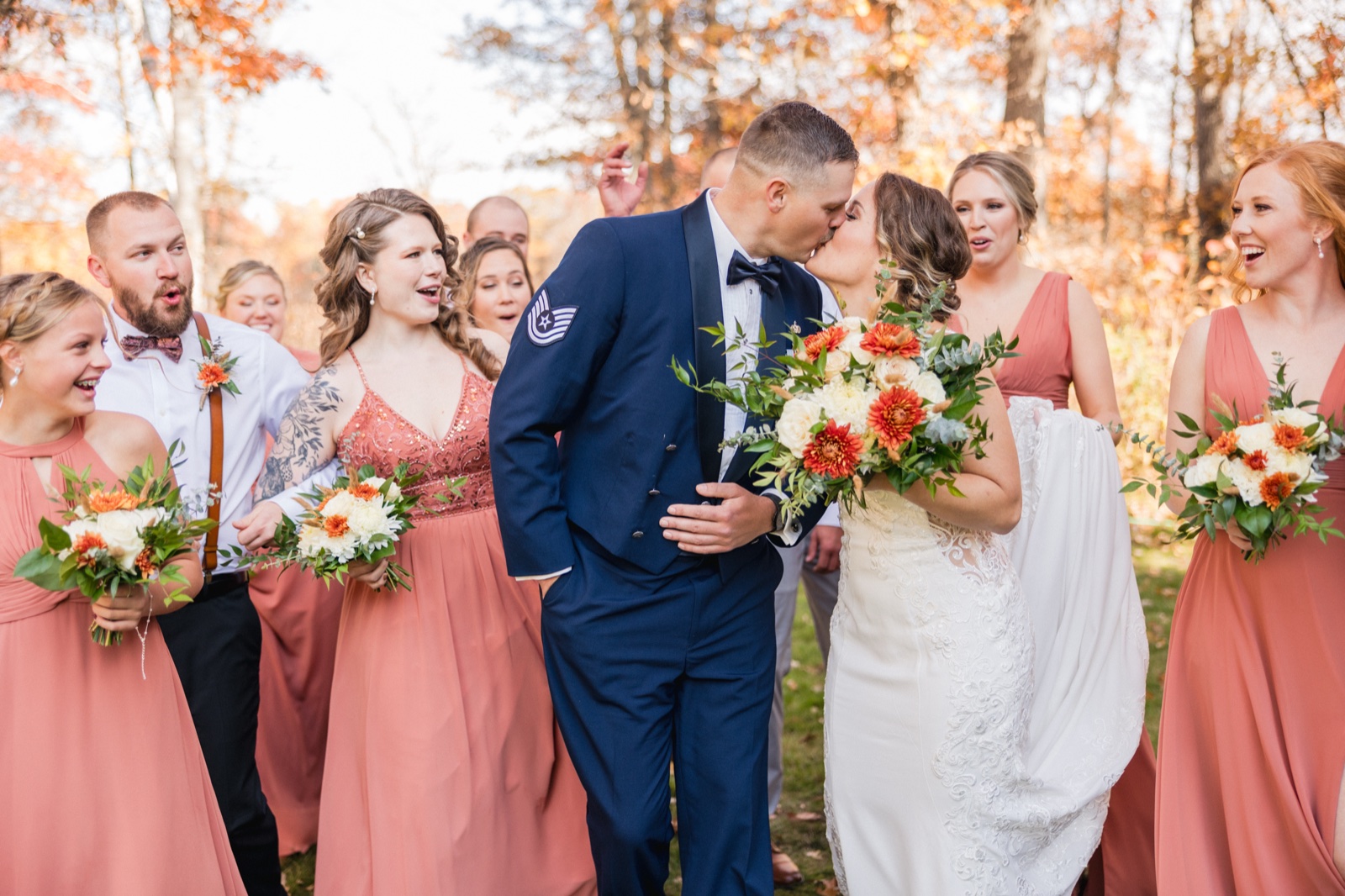 The bride and groom kiss at the center of the frame while their bridal party reacts with open-mouthed laughter and cheers, holding coral and white bouquets against autumn-lit trees — Tim Larsen Photography, Brainerd Lakes MN