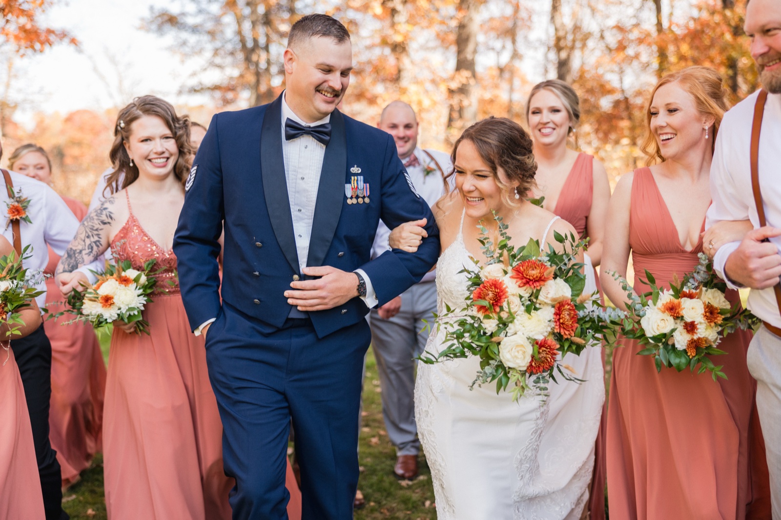 The bride and groom walk forward laughing arm-in-arm, surrounded by their bridal party in rust-colored dresses holding autumn bouquets, with backlit fall foliage behind them — Tim Larsen Photography, Brainerd Lakes MN