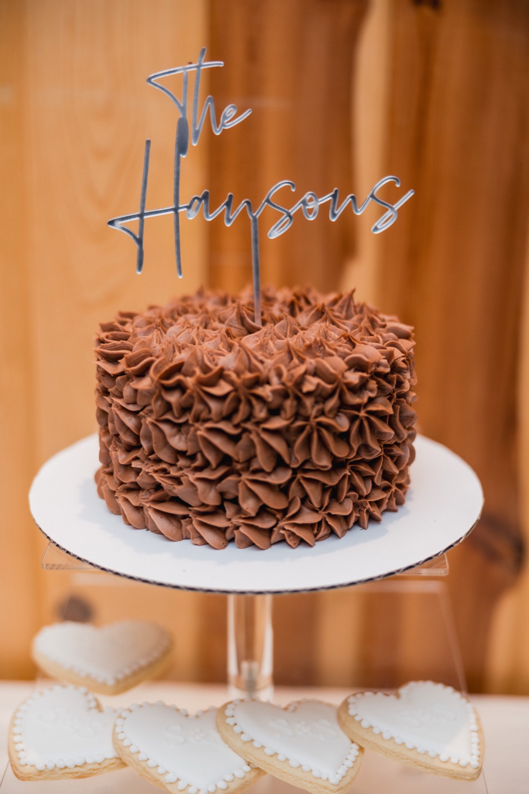 A chocolate-frosted single-tier wedding cake with a mirrored 'The Haysons' topper sits on a white stand surrounded by heart-shaped iced cookies — Tim Larsen Photography, Brainerd Lakes MN