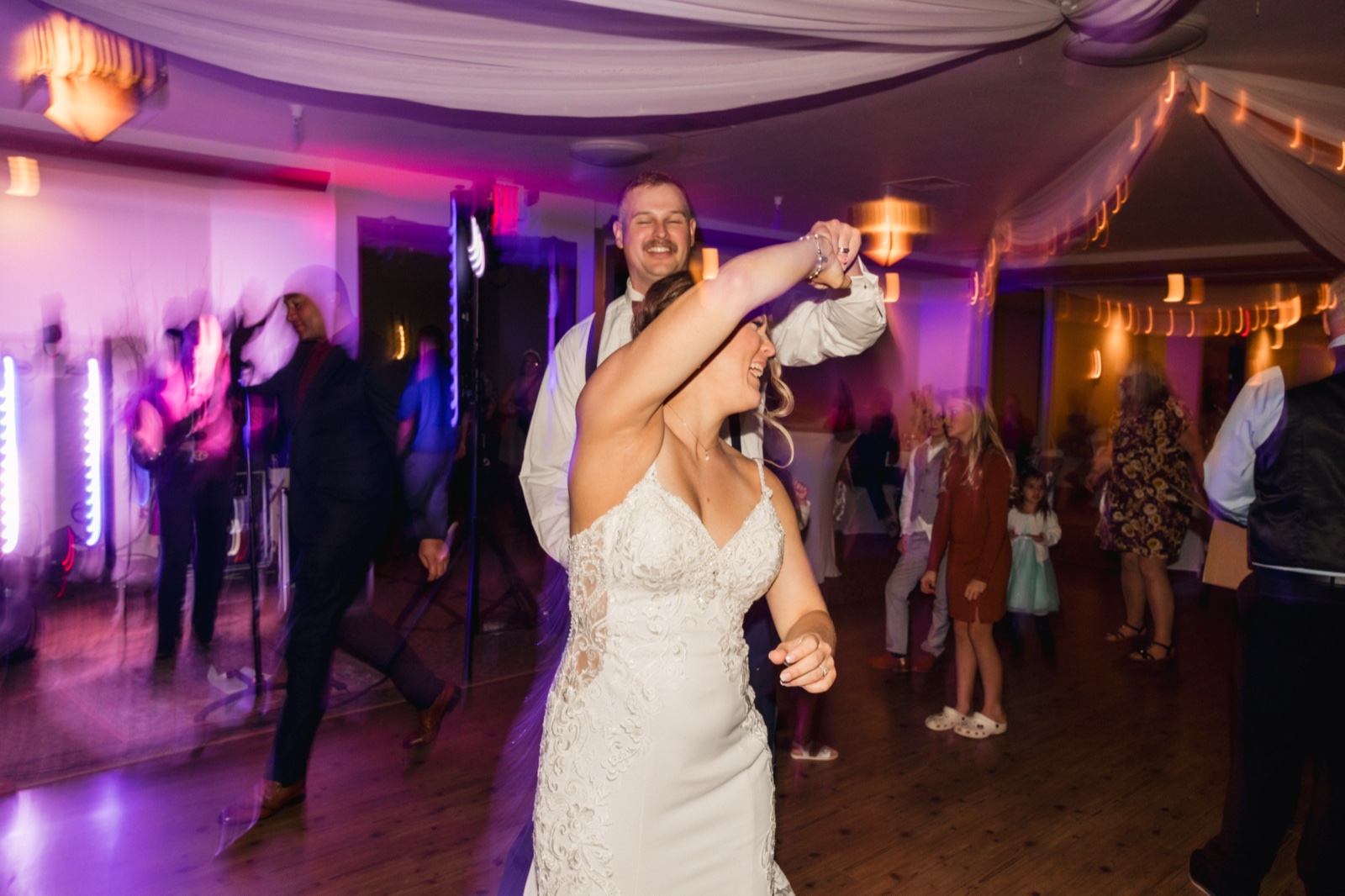 The groom spins the bride under his arm on a dance floor lit with purple uplights and draped ceiling fabric, guests dancing around them — Tim Larsen Photography, Brainerd Lakes MN