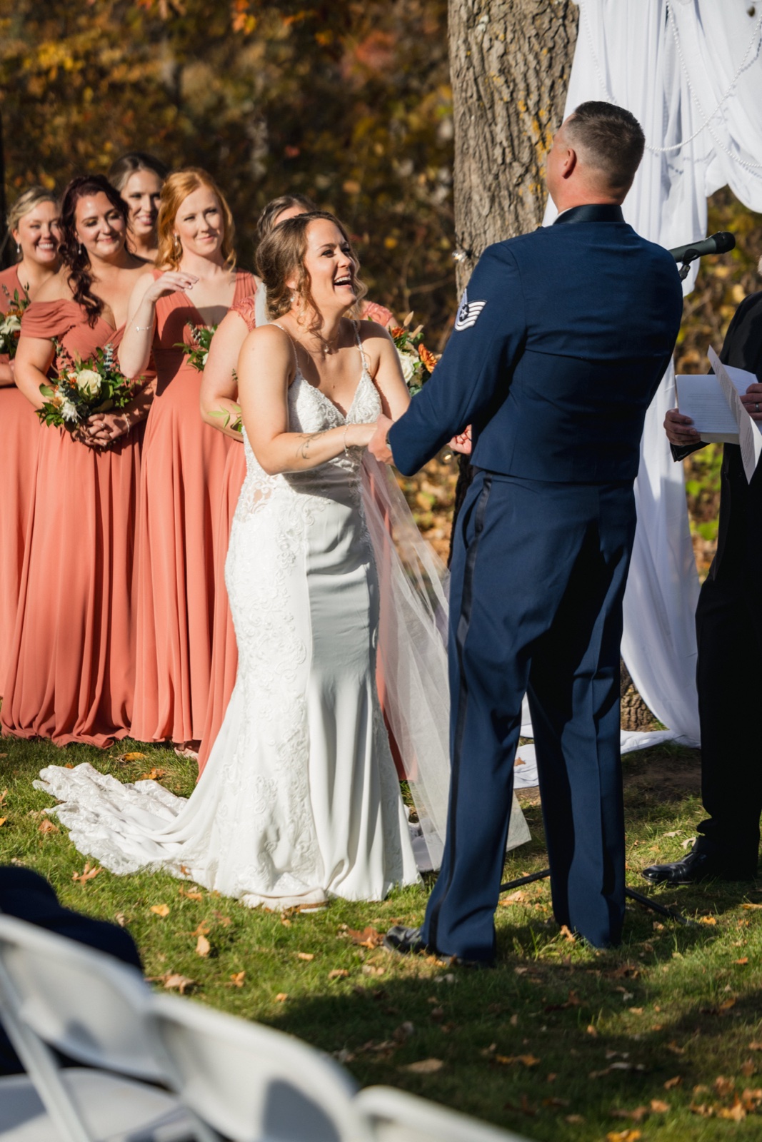 The bride laughs openly while holding hands with the groom in his Air Force dress uniform during their outdoor autumn ceremony, with bridesmaids in terracotta dresses watching from behind — Tim Larsen Photography, Brainerd Lakes MN
