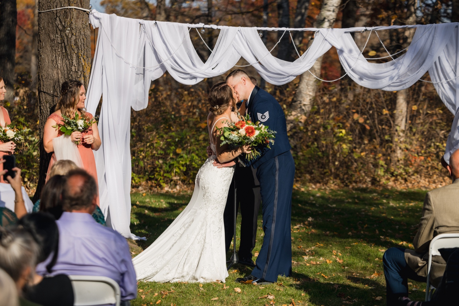 The bride and groom, in his Air Force dress uniform, share their first kiss beneath a white draped arch with autumn trees behind them — Tim Larsen Photography, Brainerd Lakes MN
