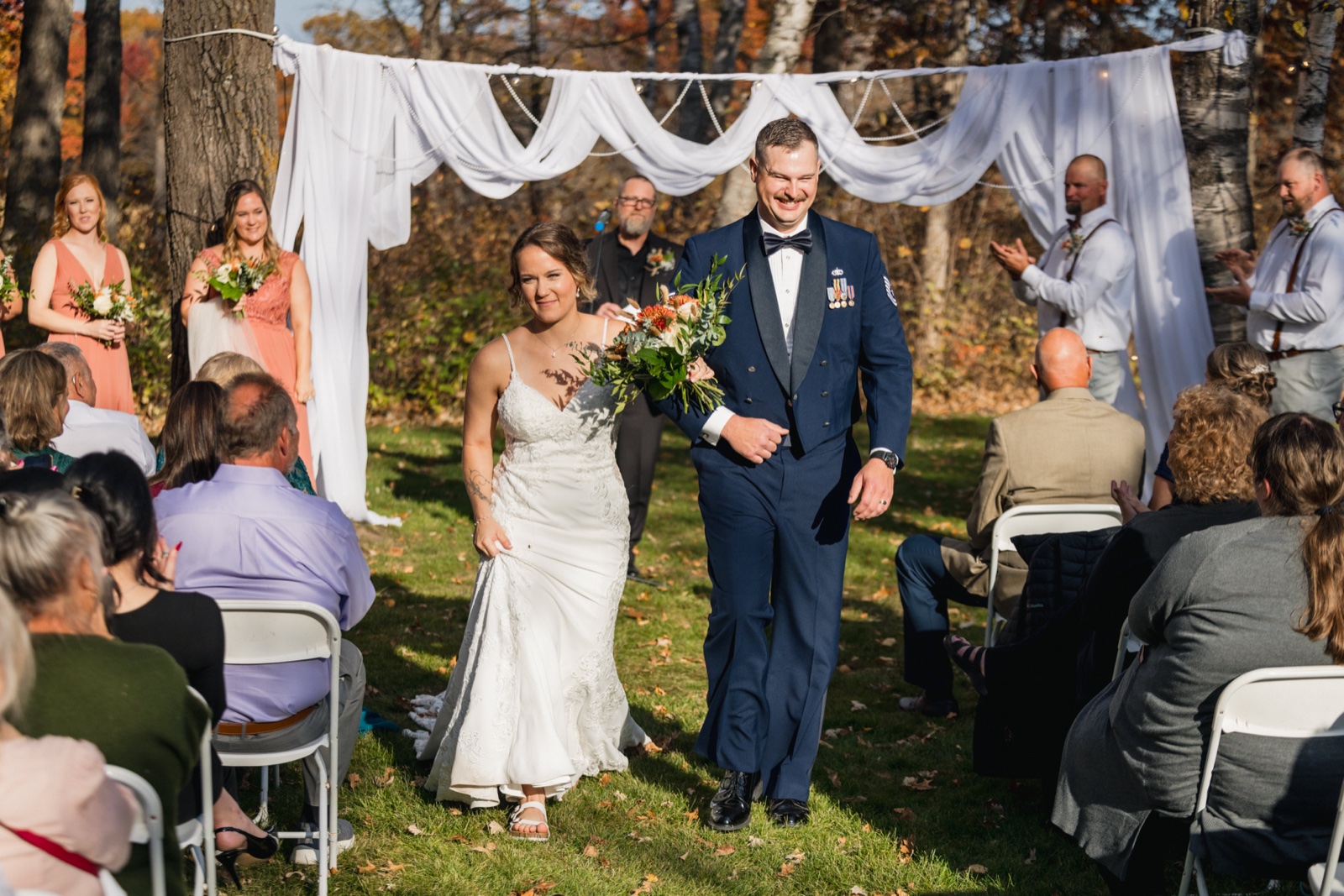 The bride and groom walk back down the aisle together after the ceremony, framed by a white draped arch and autumn trees, with guests and bridal party watching on either side — Tim Larsen Photography, Brainerd Lakes MN