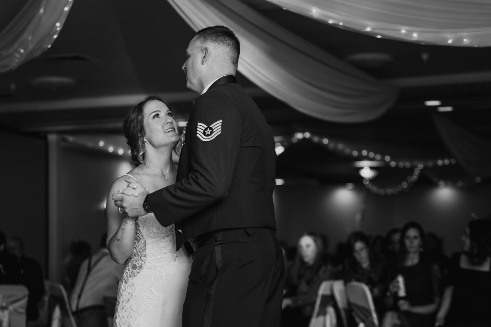 The bride looks up at her groom, who wears a military dress uniform, as they share their first dance beneath draped fabric and string lights — Tim Larsen Photography, Brainerd Lakes MN