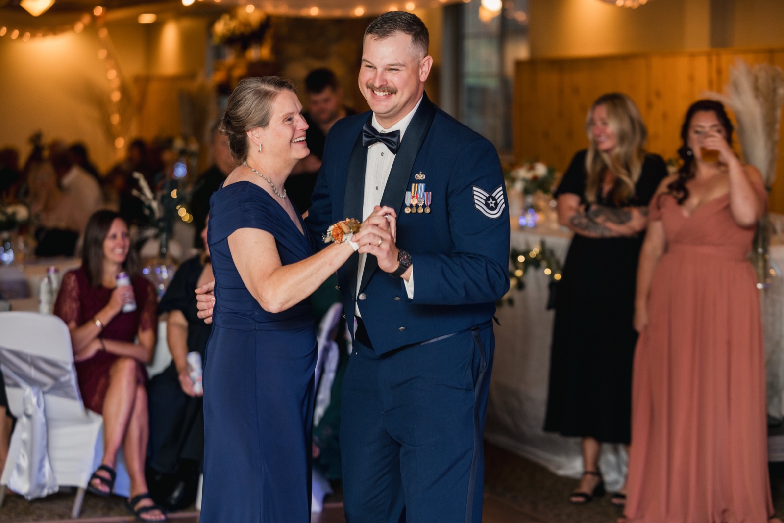 The groom in his Air Force dress blues laughs while dancing with his mother as guests watch and smile in the background — Tim Larsen Photography, Brainerd Lakes MN