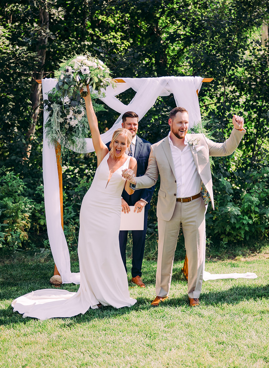 Couple celebrating after outdoor ceremony under a wooden arch with greenery — Tim Larsen Photography, Brainerd Lakes MN