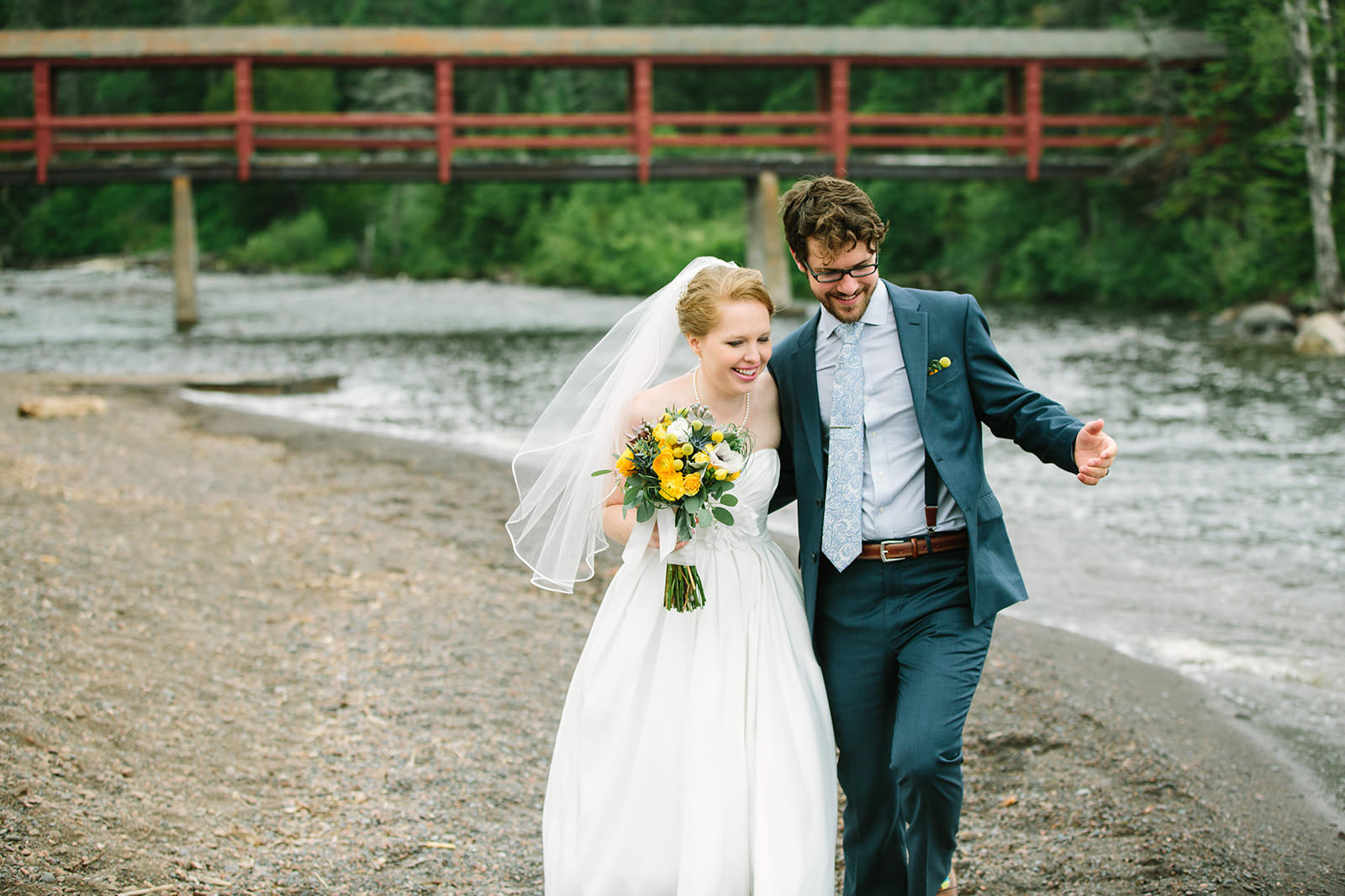 Couple laughing and walking along a river beach on the North Shore with red bridge — Tim Larsen Photography, Brainerd Lakes MN