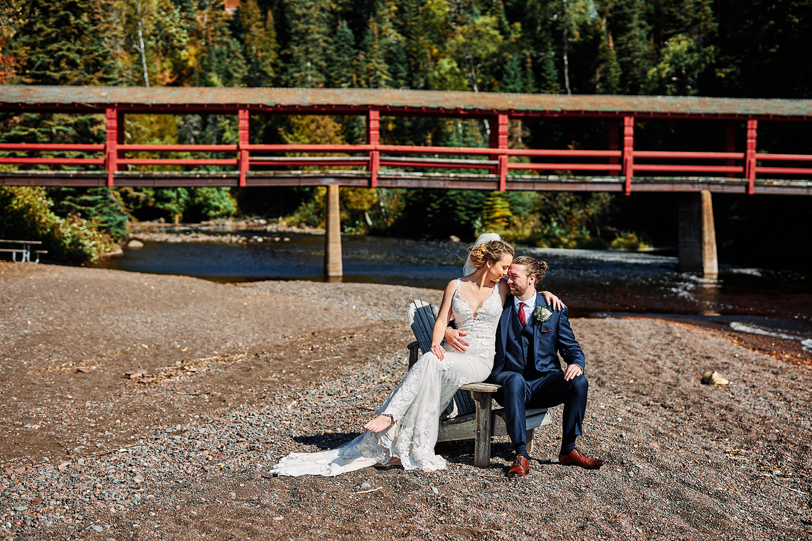 Couple sitting together on the river beach with red bridge and fall color — North Shore — Tim Larsen Photography, Brainerd Lakes MN